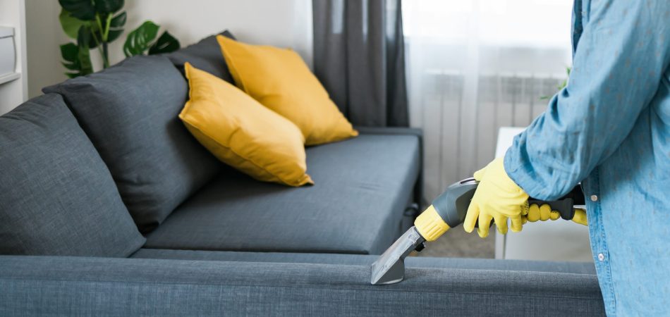 A cropped view of a man’s hands wearing white gloves using a vacuum on a sofa.